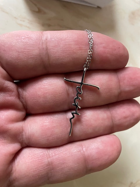 Faith Cross necklace featuring the word “faith” written vertically in script as part of the cross design, held in a hand against a light beige background.
